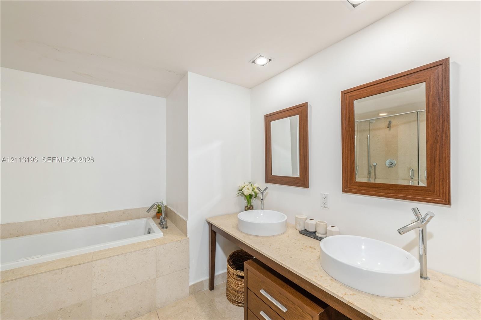 Dual vessel sinks with chrome faucets, stone-topped vanity with wood-finish cabinetry, matching wood-framed mirrors, and a tiled soaking tub