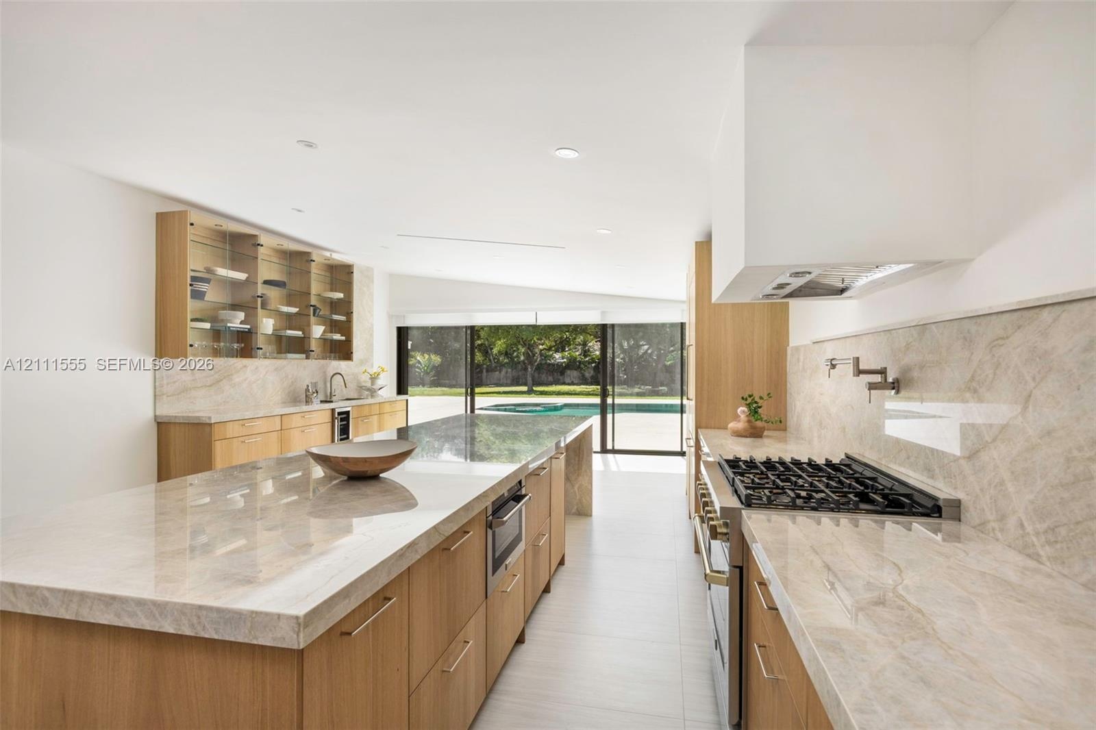 Kitchen featuring light wood-finish cabinetry, natural stone countertops, a large center island, a built-in gas cooktop with pot filler, and glass-fro