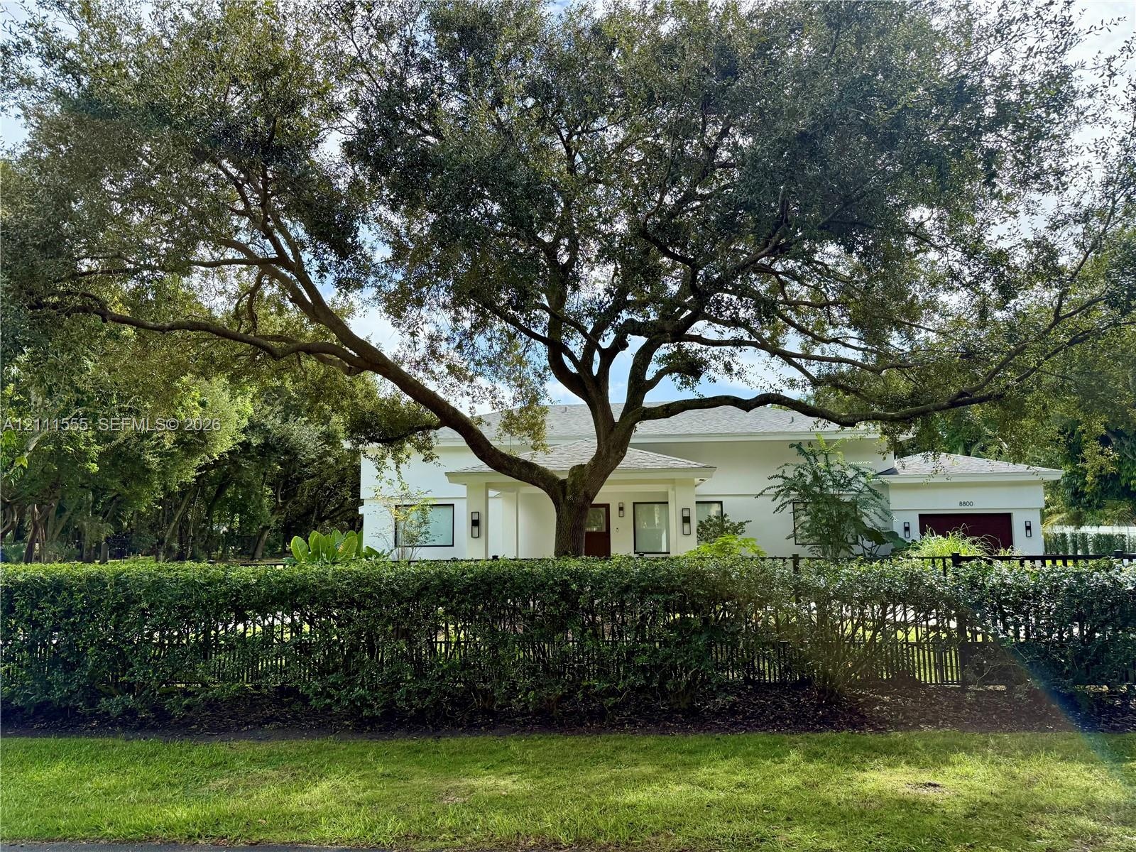 Single-story exterior featuring a light-toned stucco facade, a dark-toned multi-car garage, an expansive mature tree, and perimeter metal fencing