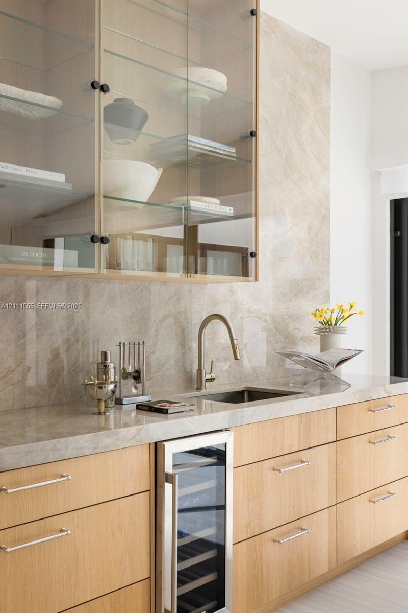 Wet bar featuring light-toned wood cabinetry, stone slab backsplash and countertop, under-counter wine refrigerator, and upper glass-front cabinetry w