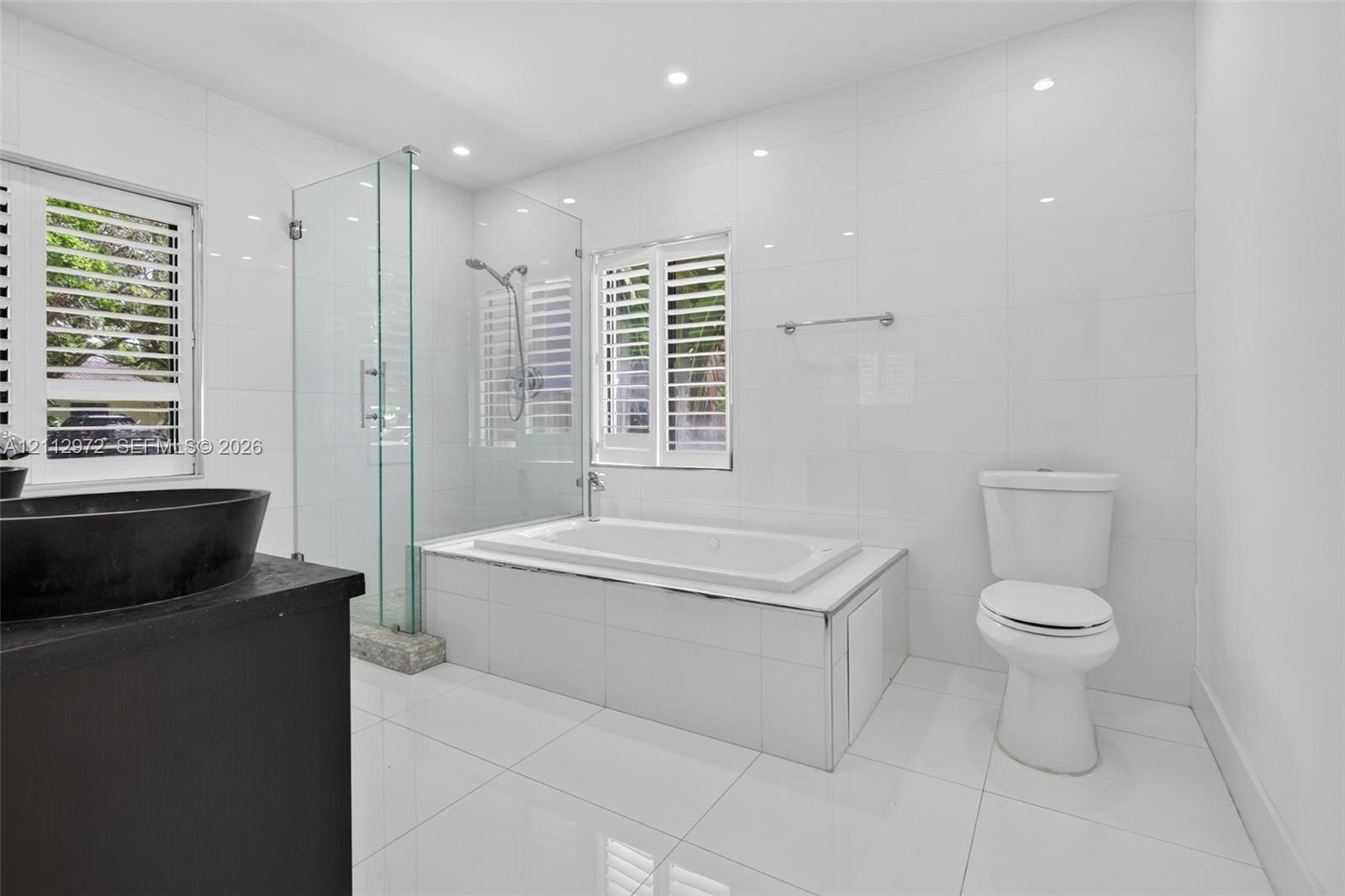 Modern bathroom featuring a glass-enclosed walk-in shower, a deep-soaking bathtub with tile surround, plantation shutters, and a dark-finish vanity wi