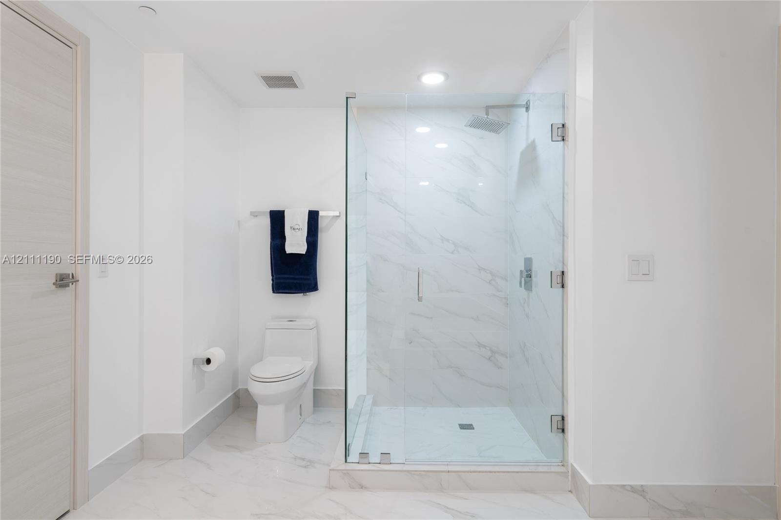 Walk-in glass-enclosed shower featuring stone-finish wall tile, dual-flush elongated toilet, and light-tone wood-finish door