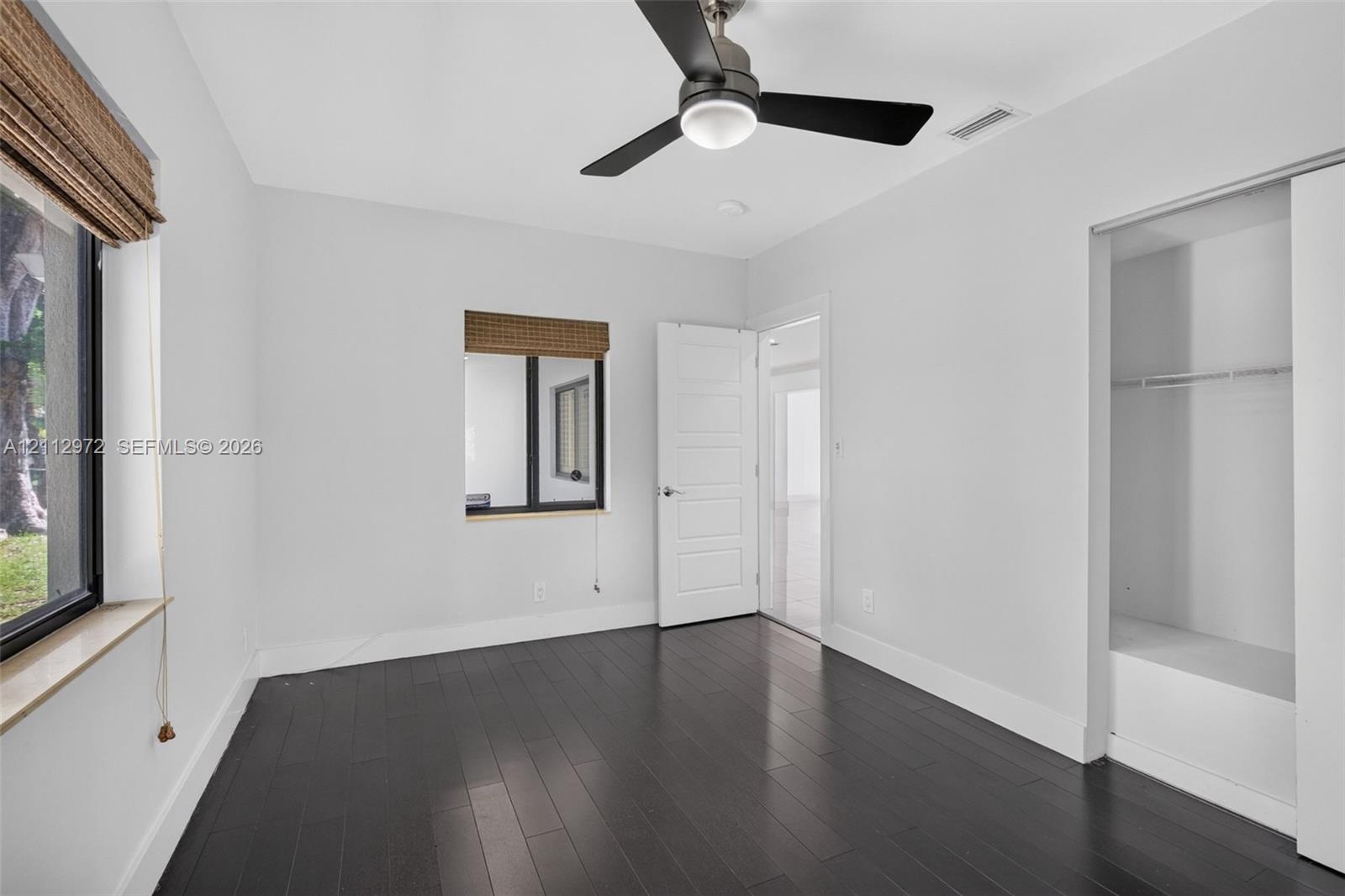 Room featuring dark wood-finish flooring, a contemporary matte black ceiling fan with integrated lighting, two windows with woven shades, and a built-