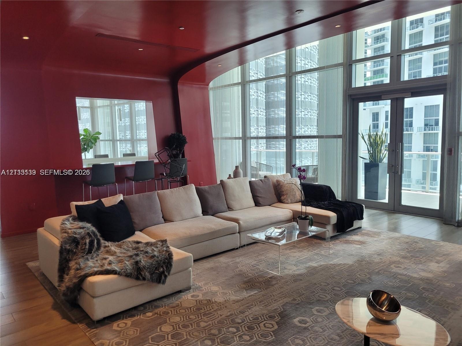 Curved, high-gloss red wall featuring an integrated service counter, floor-to-ceiling windows, recessed ceiling lighting, and double-door balcony acce