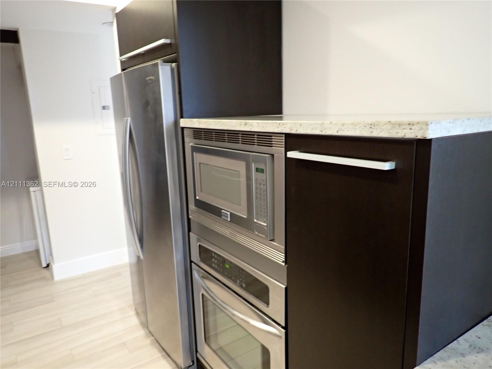 Kitchen featuring dark-finish cabinetry, stainless steel refrigerator, built-in stainless steel microwave and oven, and light-toned stone-finish count