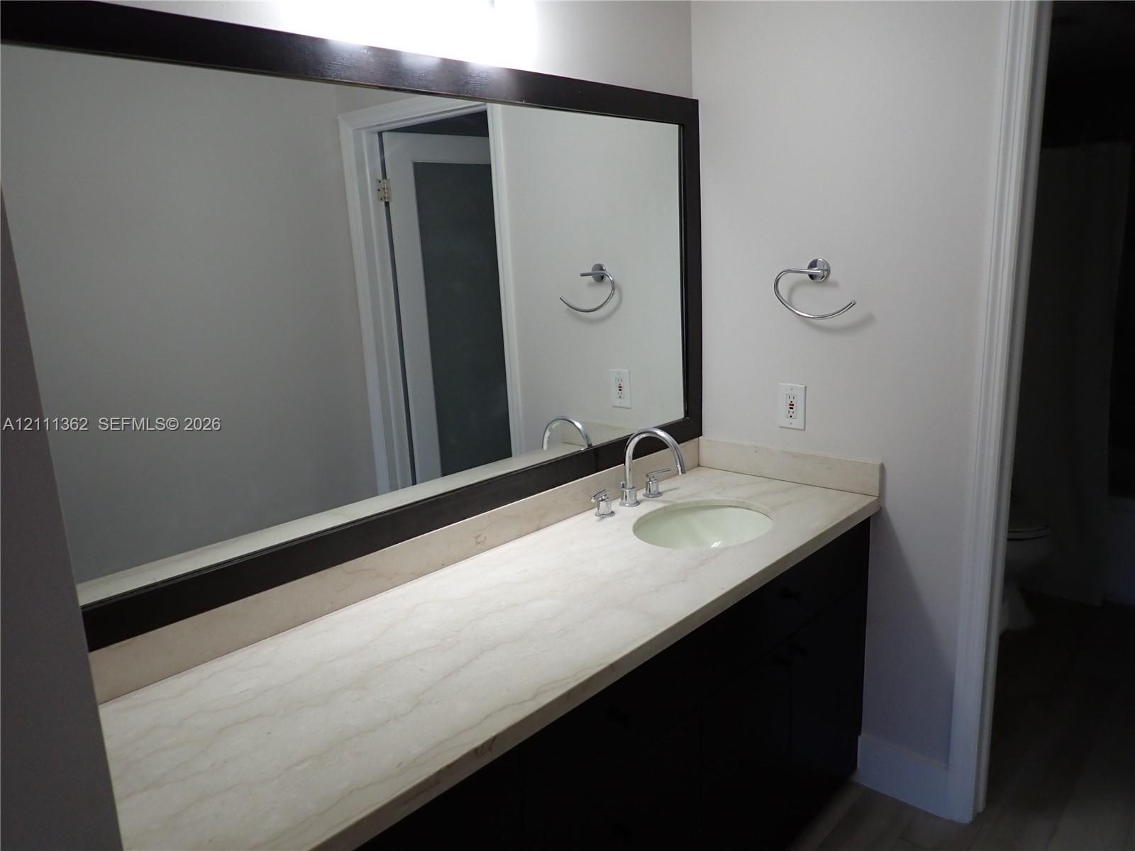 Vanity with stone-finish countertop, integrated oval sink, chrome widespread faucet, and full-width wall mirror with dark wood-finish frame