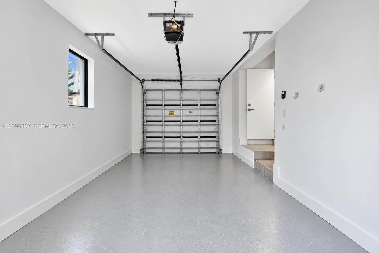 Interior garage featuring smooth white walls, a finished epoxy floor, an automatic overhead door opener, a small rectangular side window, and an inter
