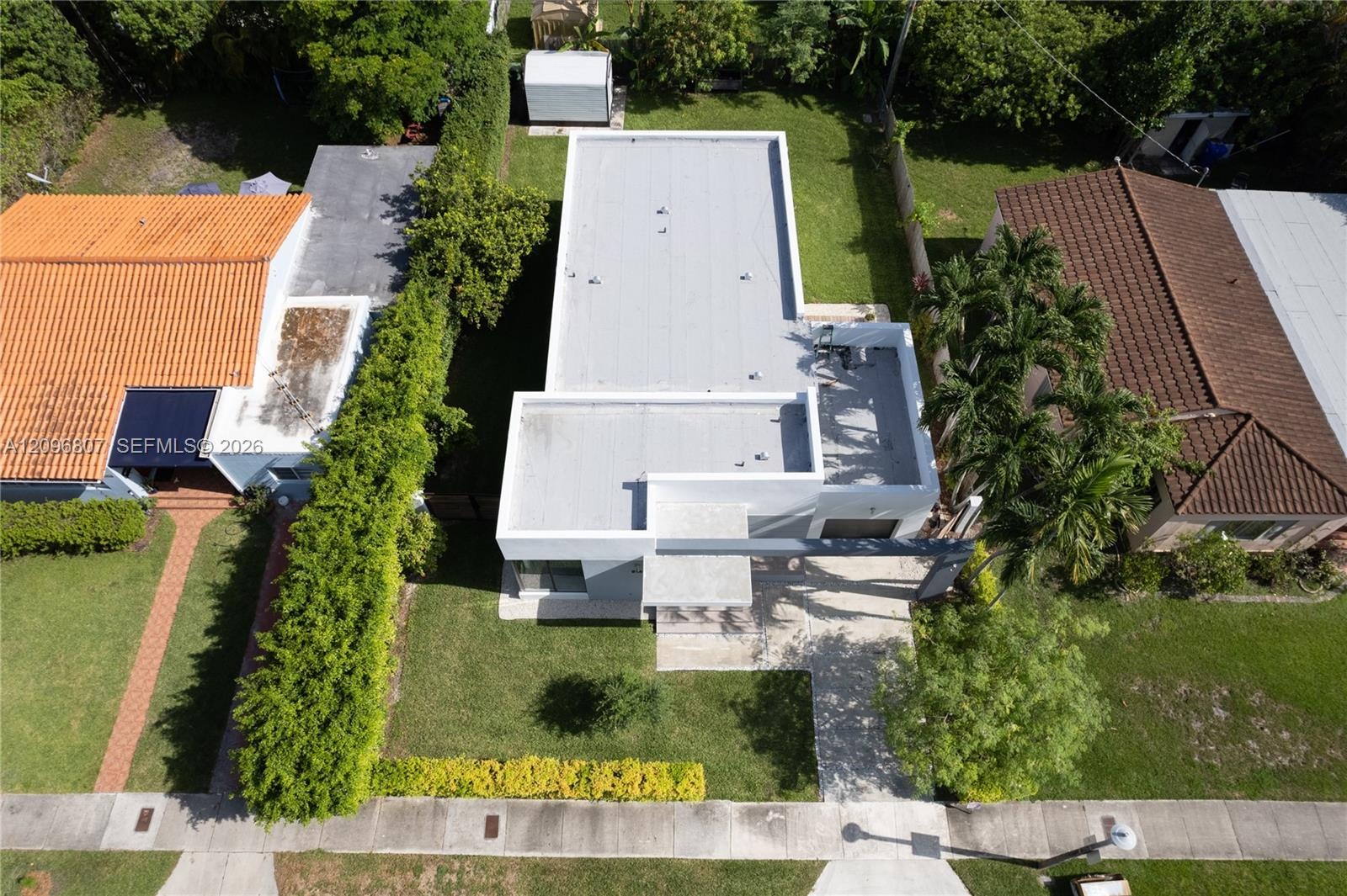 Aerial view of a flat-roof residence featuring a paved walkway, manicured lawn, established landscaping, and an enclosed storage structure