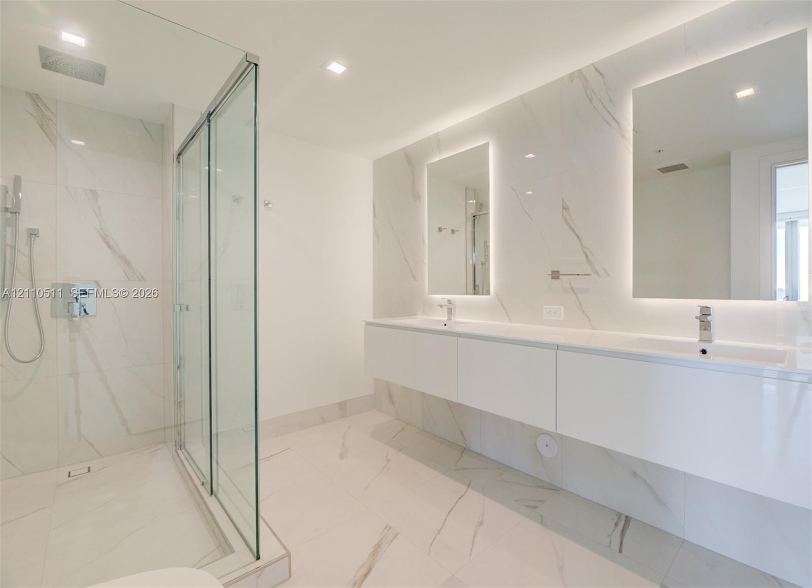 Floating dual-sink vanity with under-cabinet lighting, floor-to-ceiling stone-tiled walls, glass-enclosed walk-in shower, recessed ceiling lighting, a
