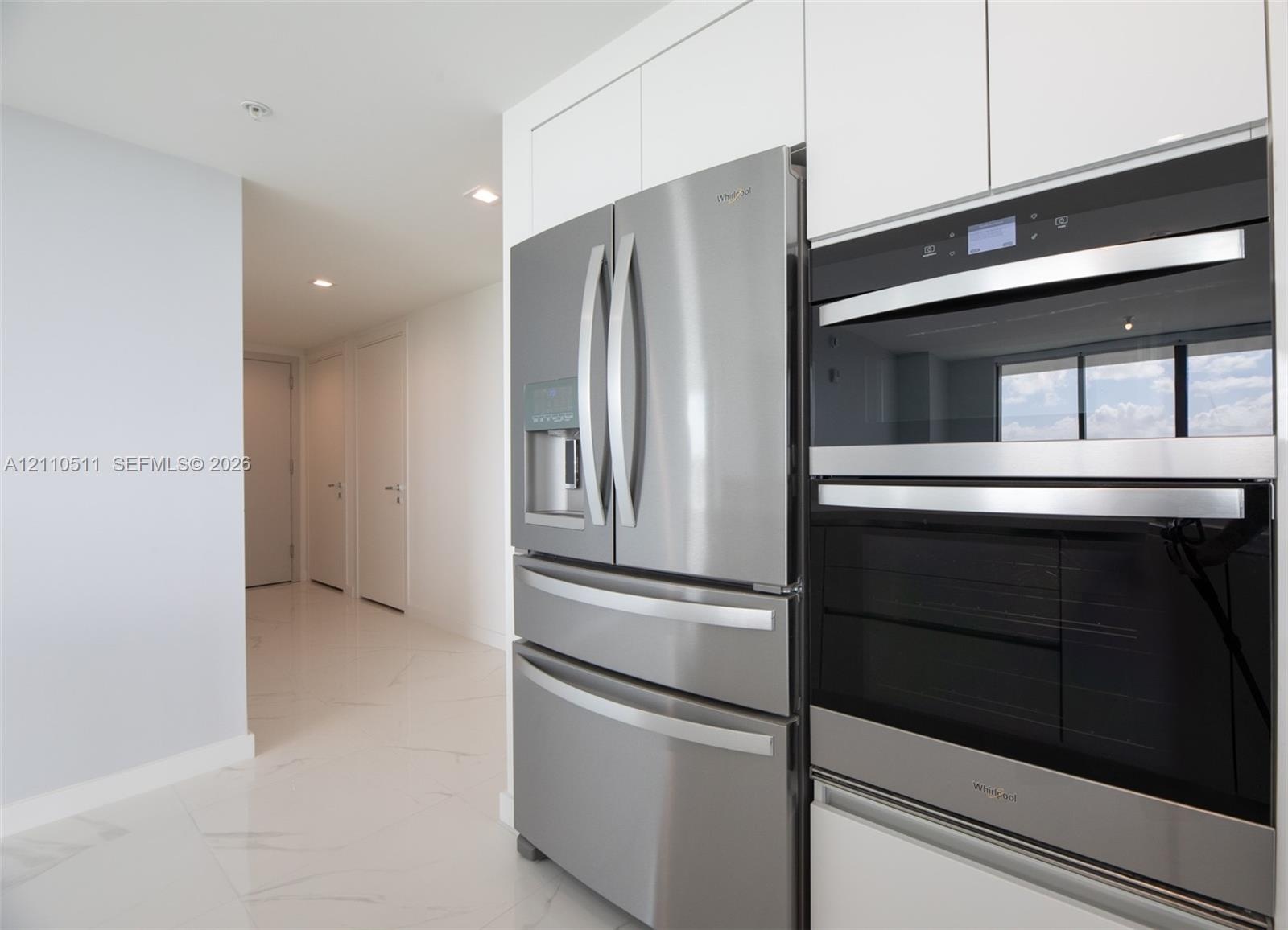 Stainless steel French door refrigerator and stacked double oven, complemented by white flat-panel cabinetry and large-format tile flooring
