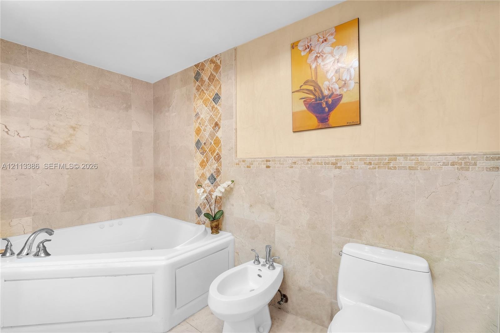 Corner soaking tub with deck-mounted chrome faucet, bidet, two-piece toilet, and floor-to-ceiling stone tile walls with decorative glass and stone mos