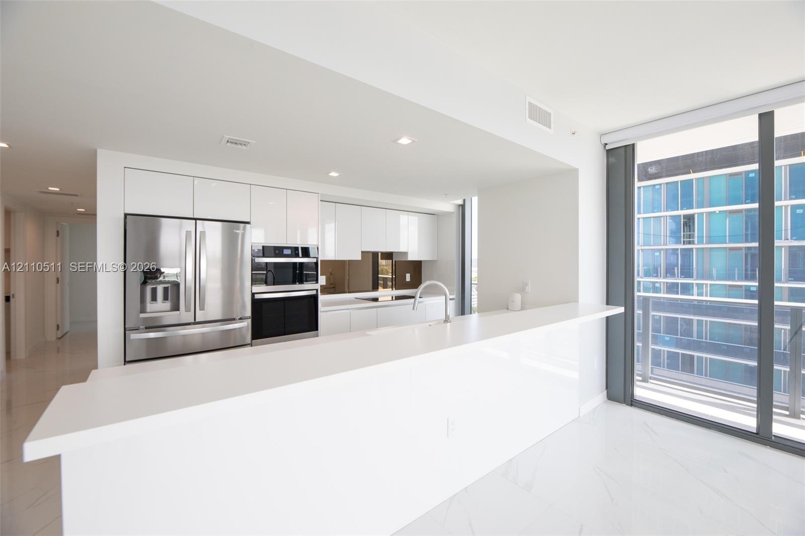 Modern kitchen featuring high-gloss white cabinetry, expansive stone countertop, stainless steel refrigerator, built-in wall oven, and floor-to-ceilin