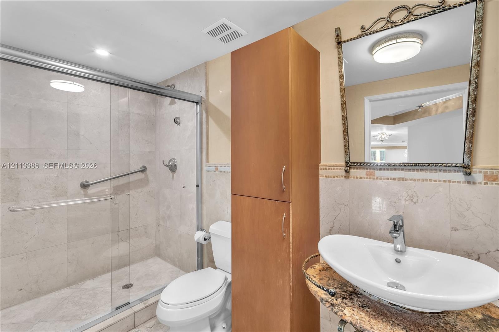 Walk-in shower with sliding glass enclosure and tiled walls, stone-finish countertop with vessel sink, tall built-in wooden cabinetry, and decorative 
