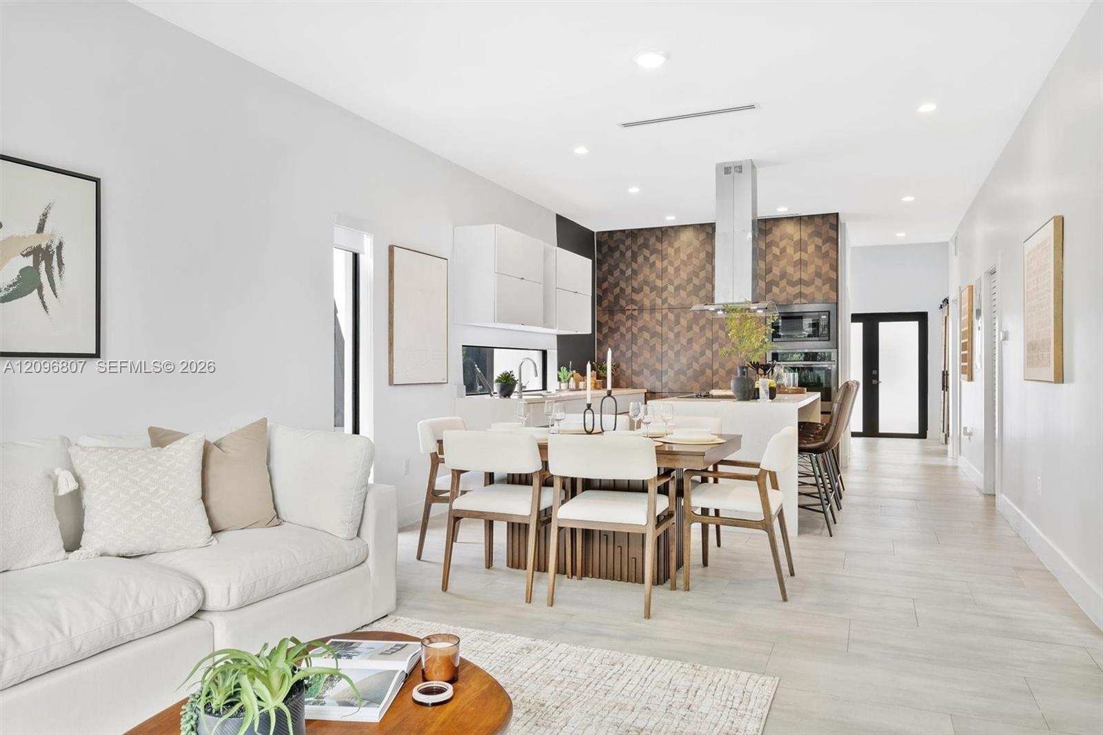 Open-concept kitchen featuring chevron-patterned backsplash tile, center island with stone countertop, stainless steel range hood, built-in wall oven,