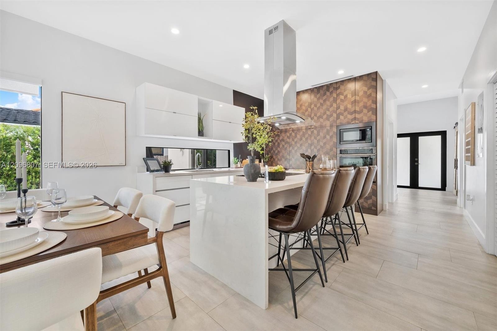 Kitchen featuring a waterfall-edge island with stone-finish countertop, high-gloss cabinetry, stainless steel range hood, patterned tile backsplash, a