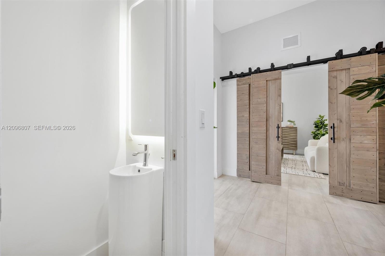 Pedestal sink with a single-handle chrome faucet, LED-backlit mirror, large-format tile flooring, and wood-finish sliding barn doors with matte black 