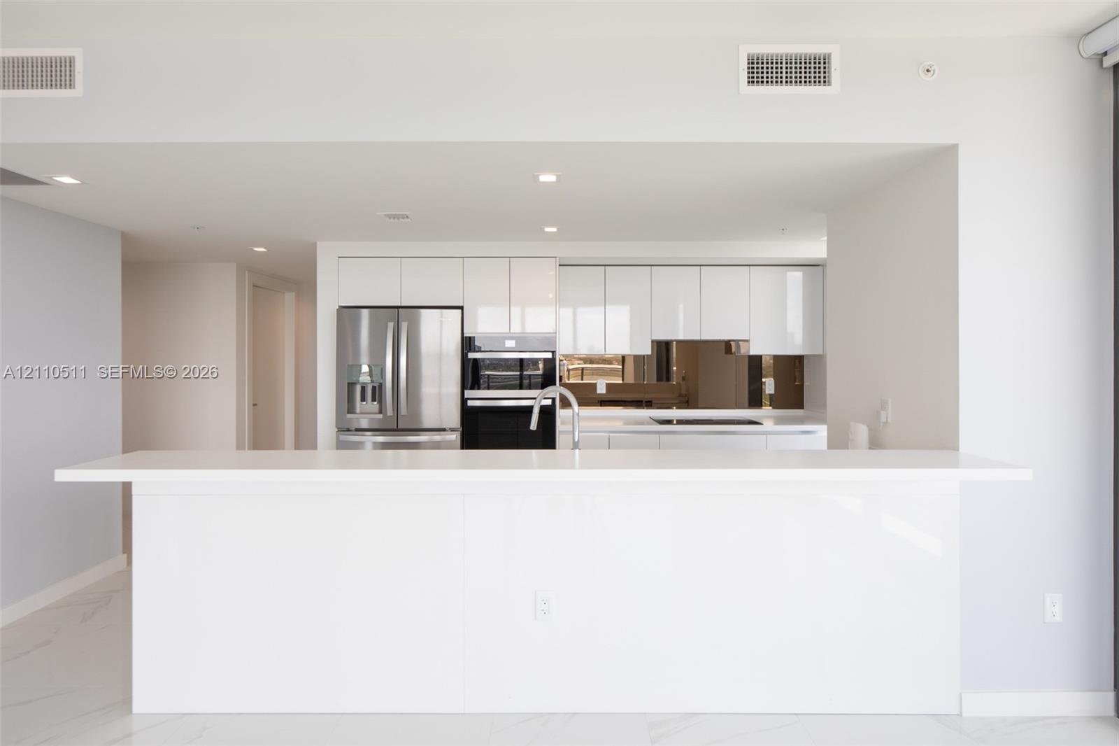 Modern kitchen featuring high-gloss white cabinetry, expansive stone-finish peninsula, stainless steel french-door refrigerator, integrated double ove