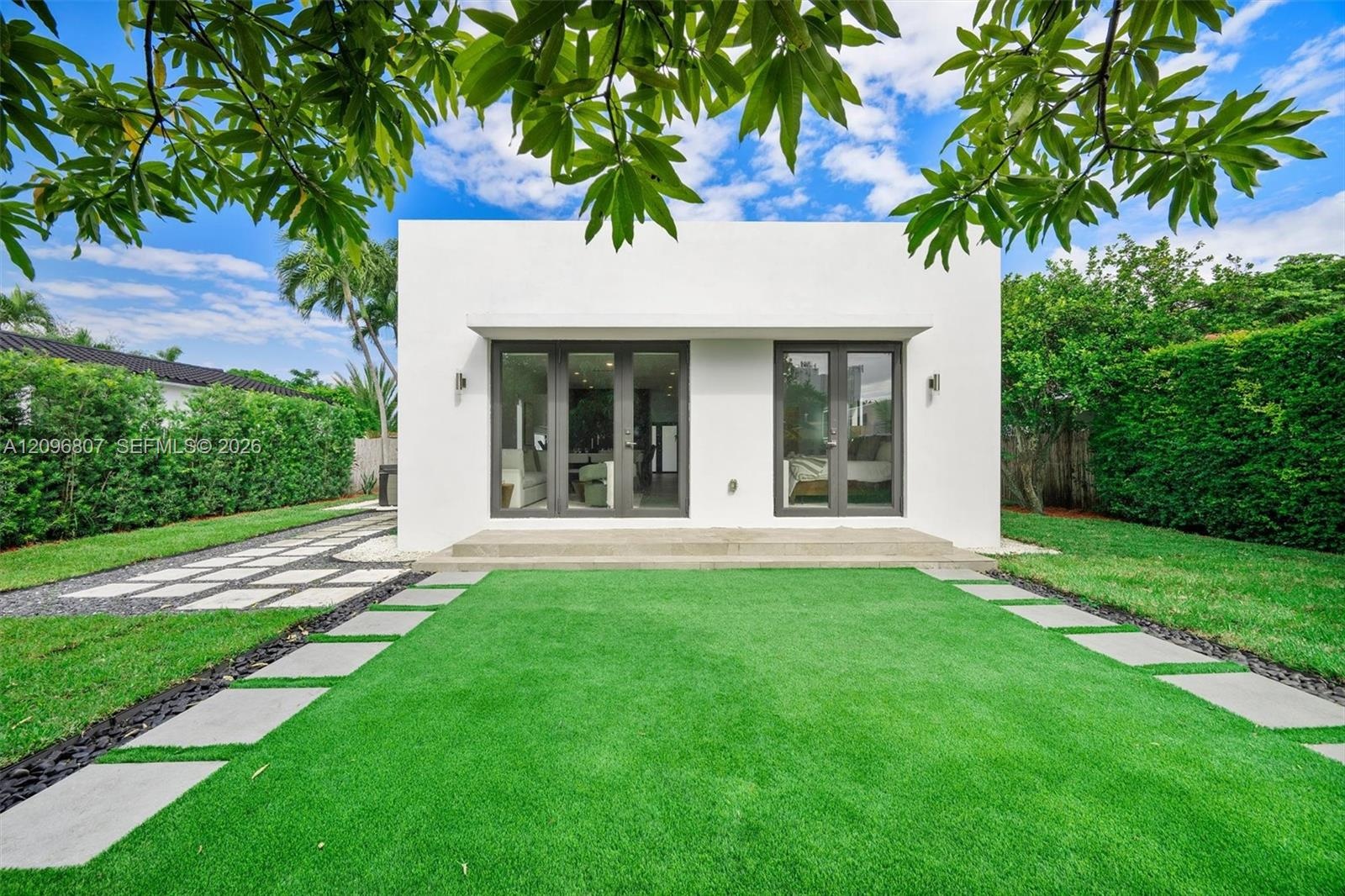 Minimalist white exterior featuring dual sets of glass-paned French doors, concrete step entry, synthetic turf lawn, and geometric stone pavers