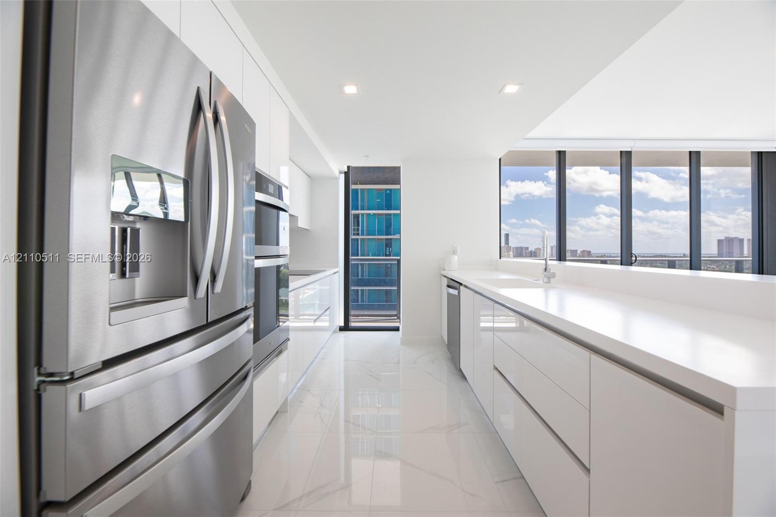 Modern kitchen featuring high-gloss white cabinetry, expansive stone countertops, stainless steel appliances, floor-to-ceiling windows, and large-form