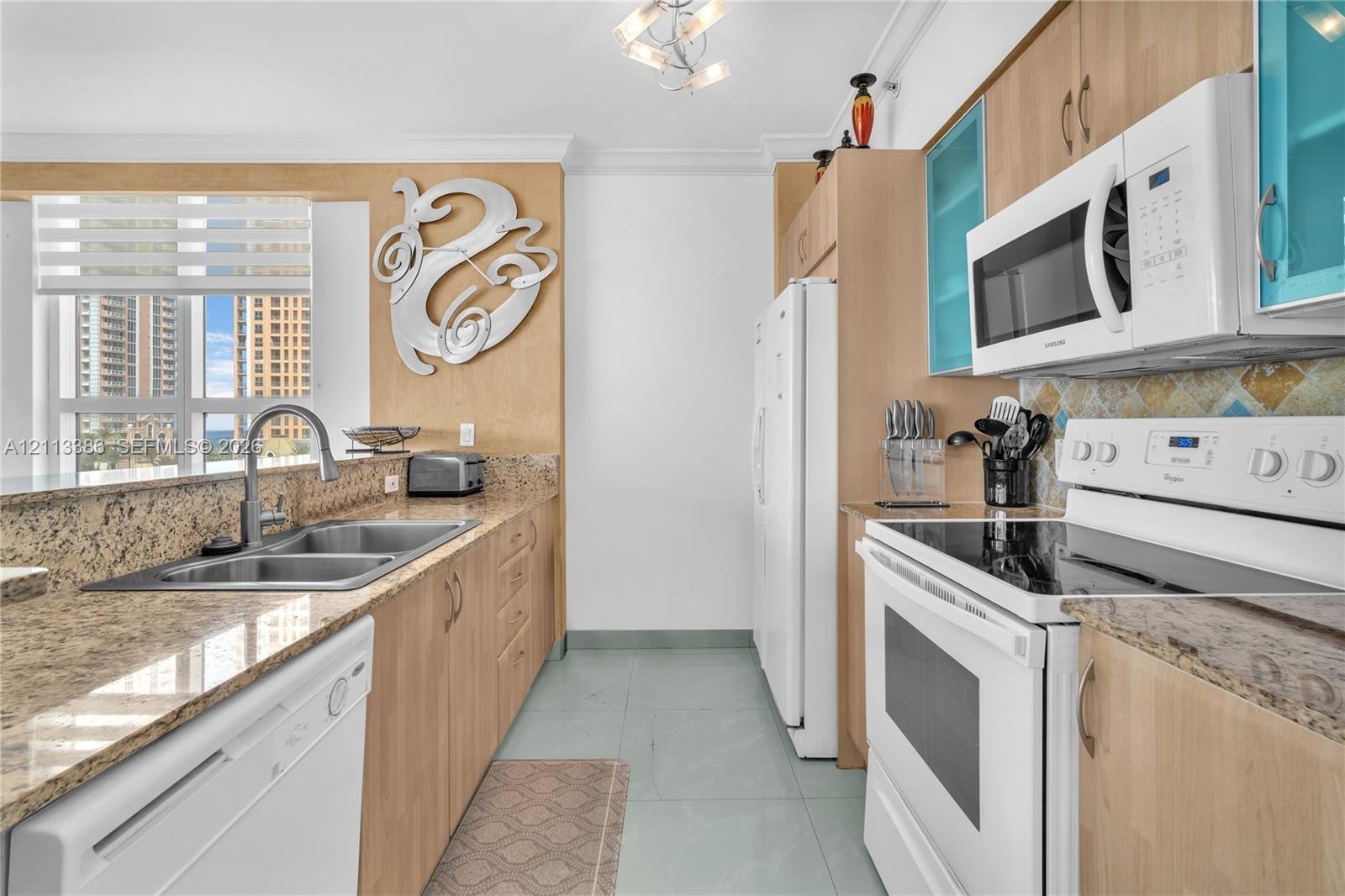 Kitchen featuring light wood-finish cabinetry, stone-surface countertops, dual-basin stainless steel sink, tile flooring, and integrated white applian