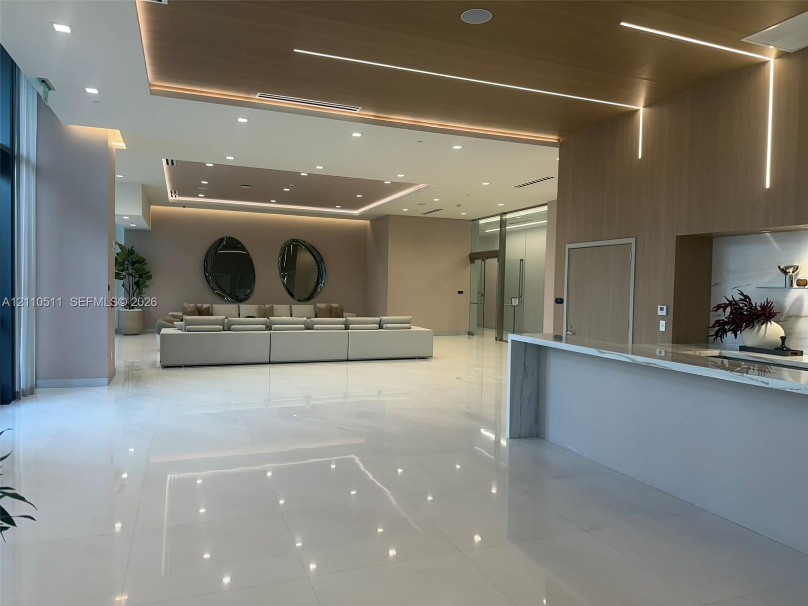 Open-concept interior featuring large-format polished tile flooring, recessed cove lighting, wood-paneled ceiling accents, and a built-in stone-top se