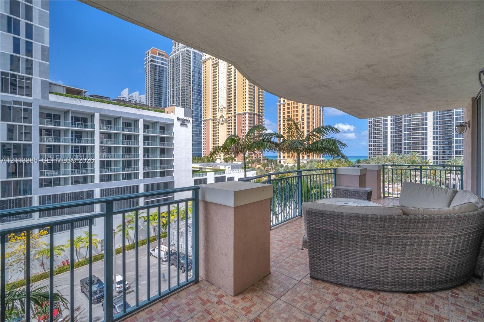 Covered balcony featuring tile flooring and metal railings, overlooking urban surroundings with palm trees and ocean glimpses