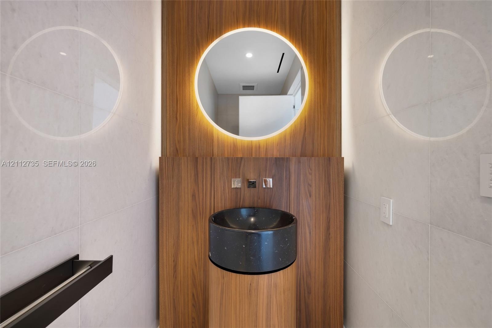Wall-mounted wood-finish paneling, circular backlit mirror, cylindrical wood-finish pedestal vanity, black stone vessel sink, and wall-mounted metalli