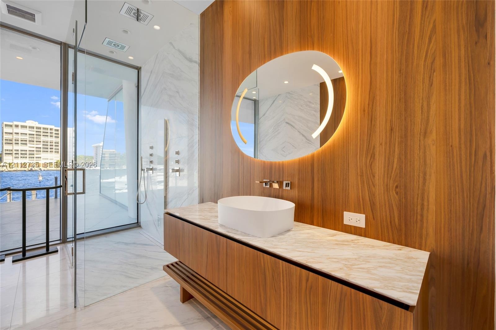Floating wood-finish vanity with a stone-top surface and vessel sink, illuminated oval mirror, floor-to-ceiling glass-enclosed shower, and polished st