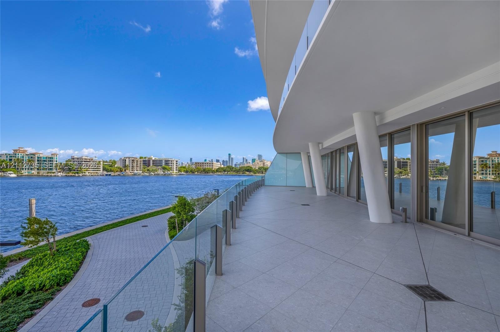 Expansive covered balcony featuring large-format tile flooring, structural support columns, full-height glass sliding doors, and a frameless glass rai