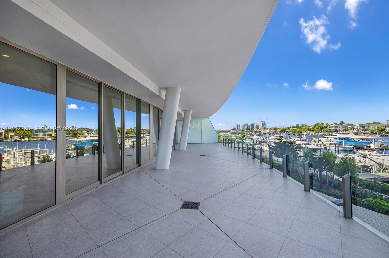 Expansive covered terrace featuring large-format tile flooring, structural support columns, floor-to-ceiling glass wall panels, and a frameless glass 