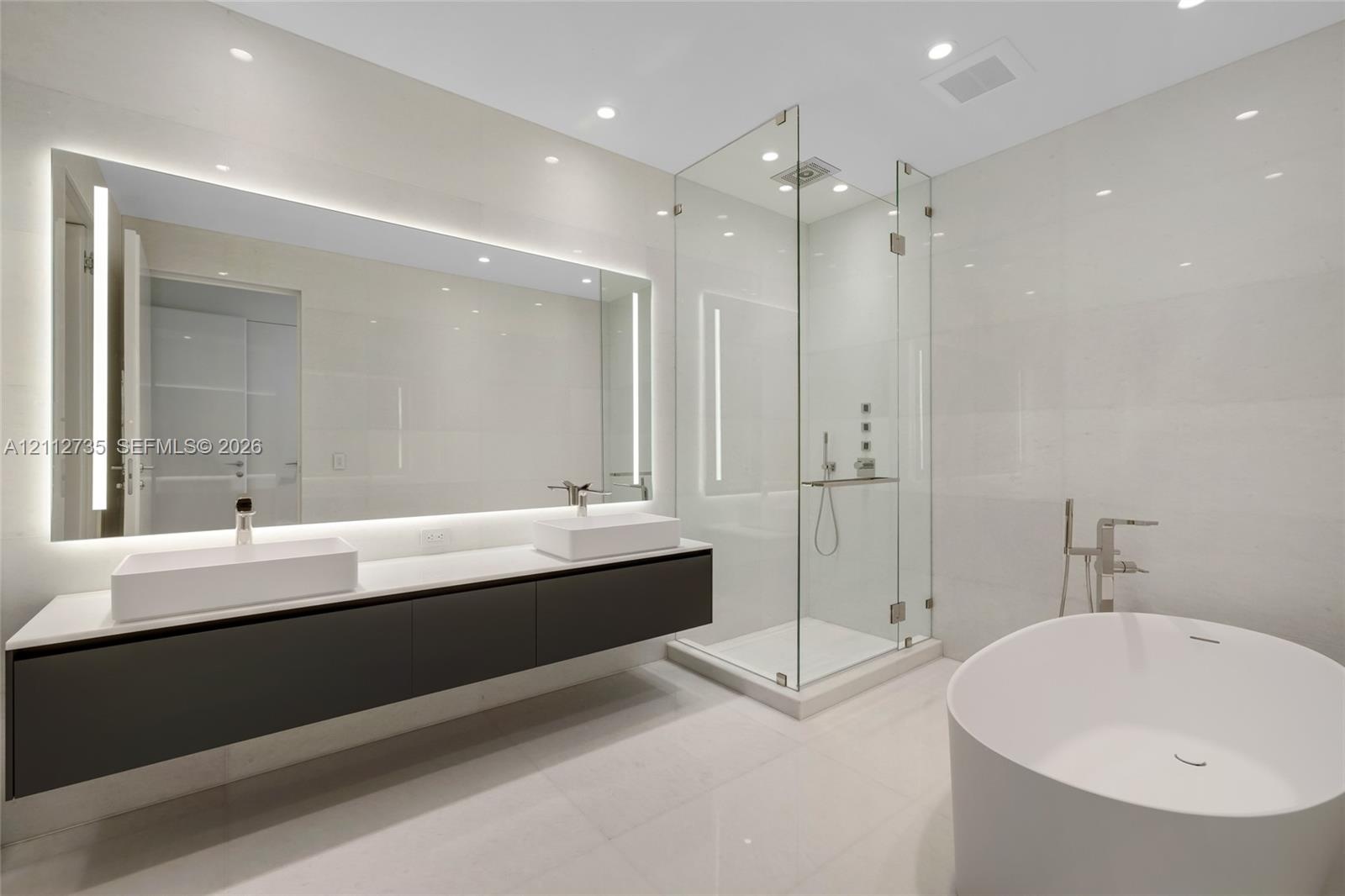 Floating double-vanity with vessel sinks, expansive LED-backlit mirror, glass-enclosed walk-in shower, and freestanding soaking tub