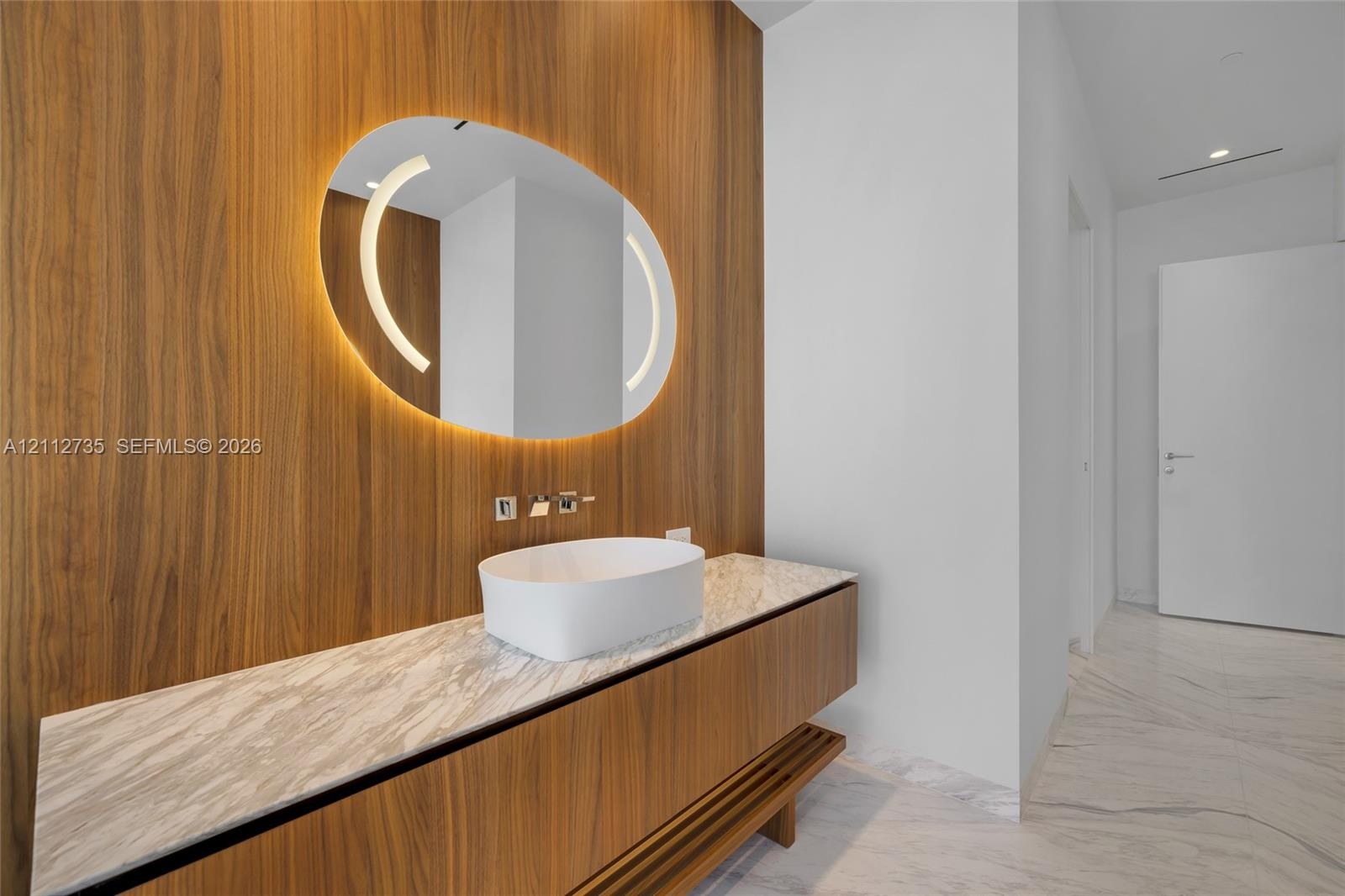 Floating wood-finish vanity with a stone countertop, white vessel sink, wall-mounted faucet, and oval backlit mirror set against a floor-to-ceiling wo