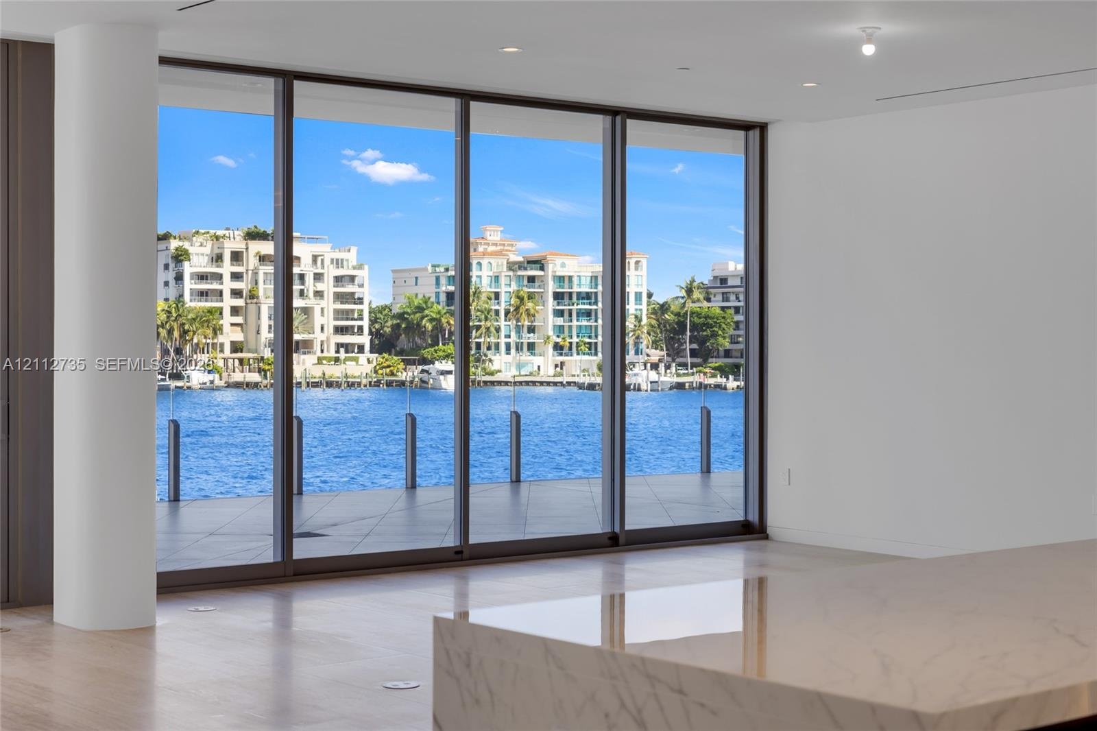 Floor-to-ceiling multi-panel glass doors, stone-topped kitchen island, polished light-toned flooring, and water frontage with glass railing