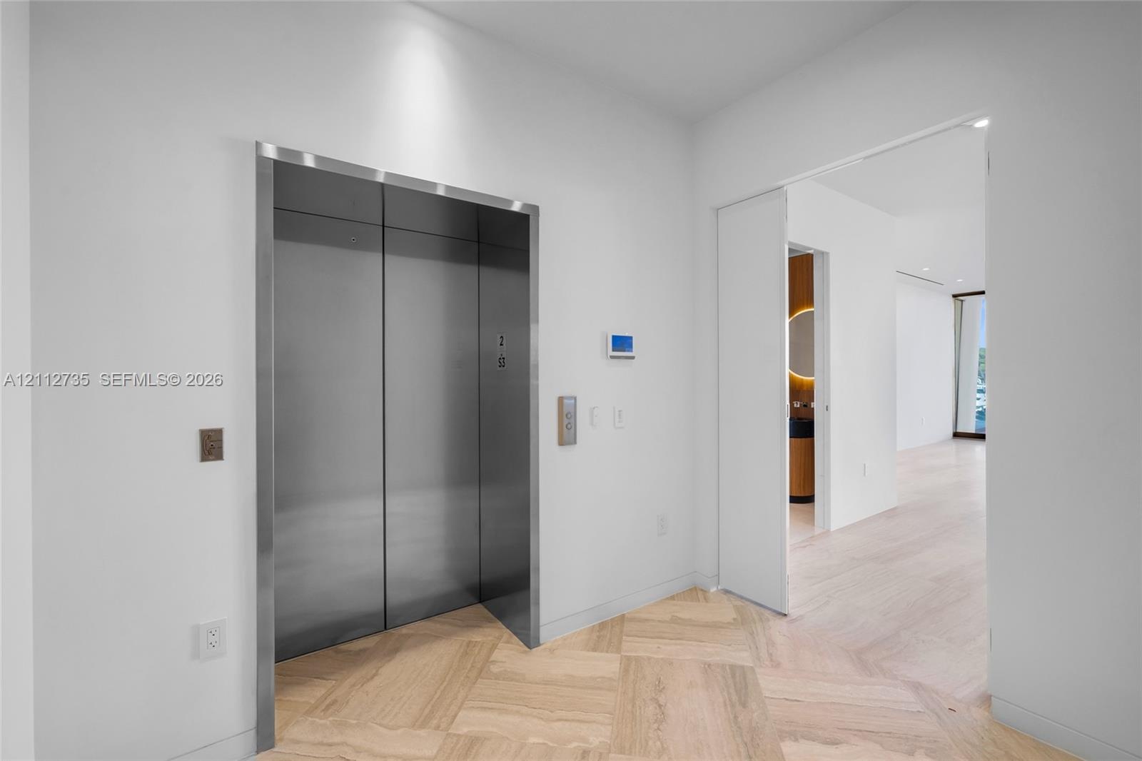 Stainless steel elevator with brushed metal finish, herringbone wood-finish flooring, and recessed ceiling lighting
