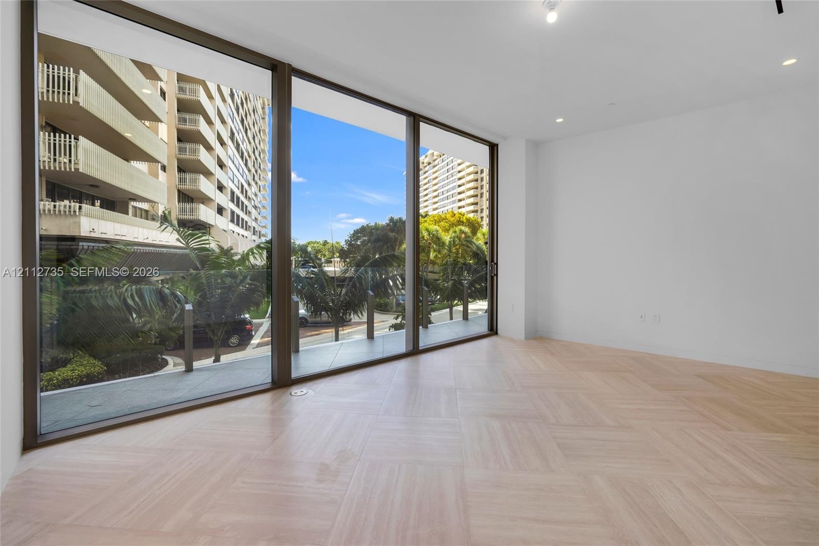 Floor-to-ceiling glass panel windows, light wood-finish herringbone flooring, recessed ceiling lighting, and a glass-railed balcony