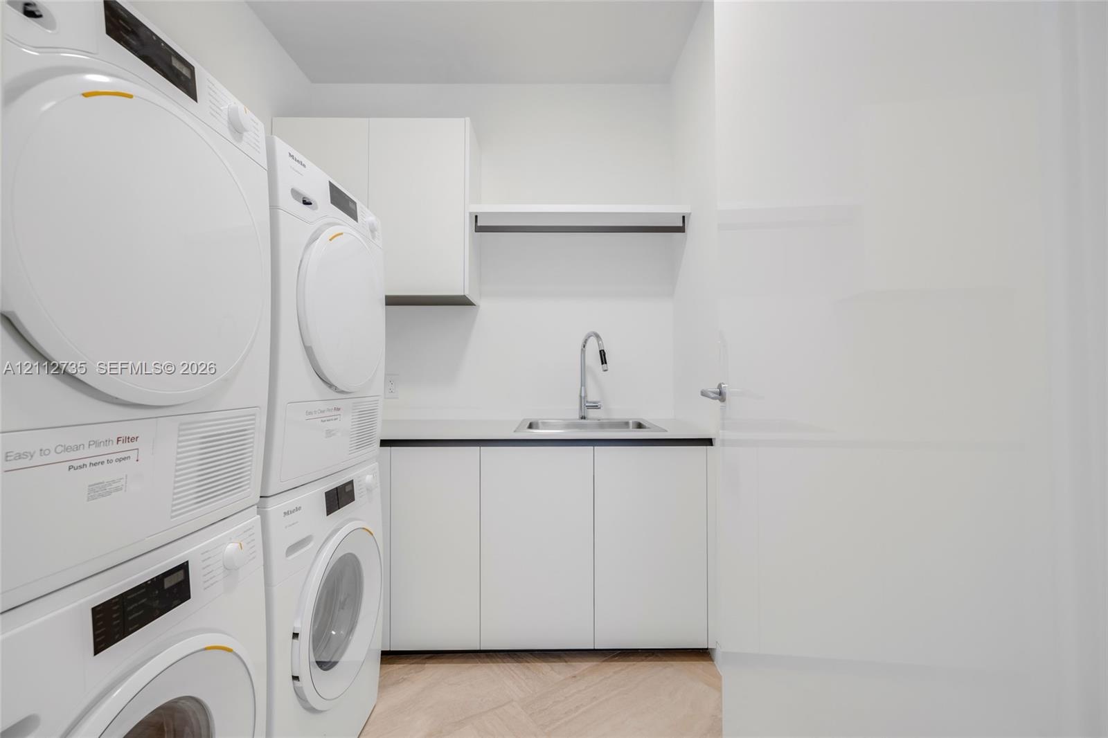 Utility area featuring stacked laundry appliances, white built-in cabinetry, a rectangular stainless steel sink with a high-arch pull-down faucet, and