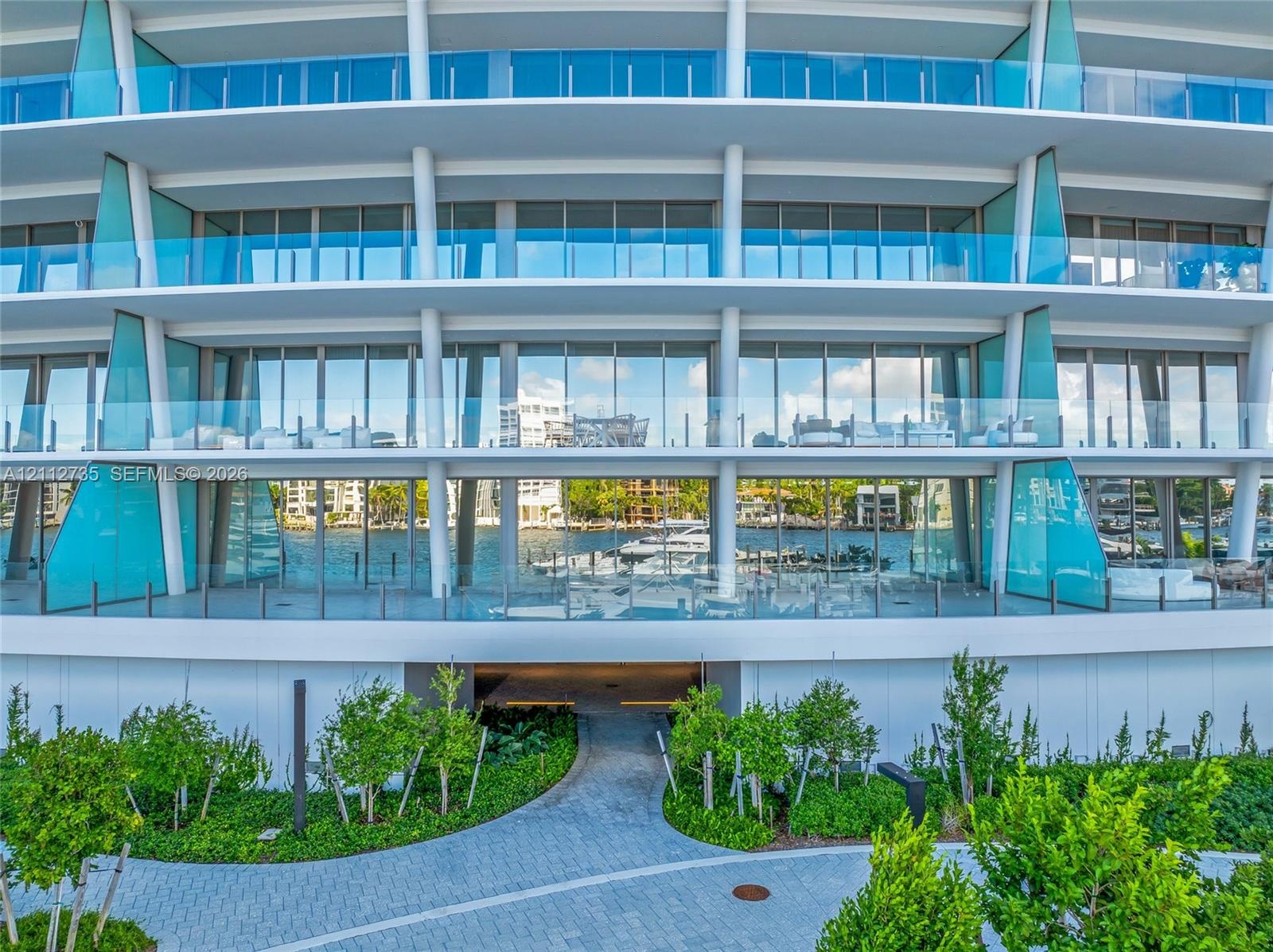 Contemporary multi-story facade featuring floor-to-ceiling glass windows, wraparound balconies with glass railings, structural white pillars, and land