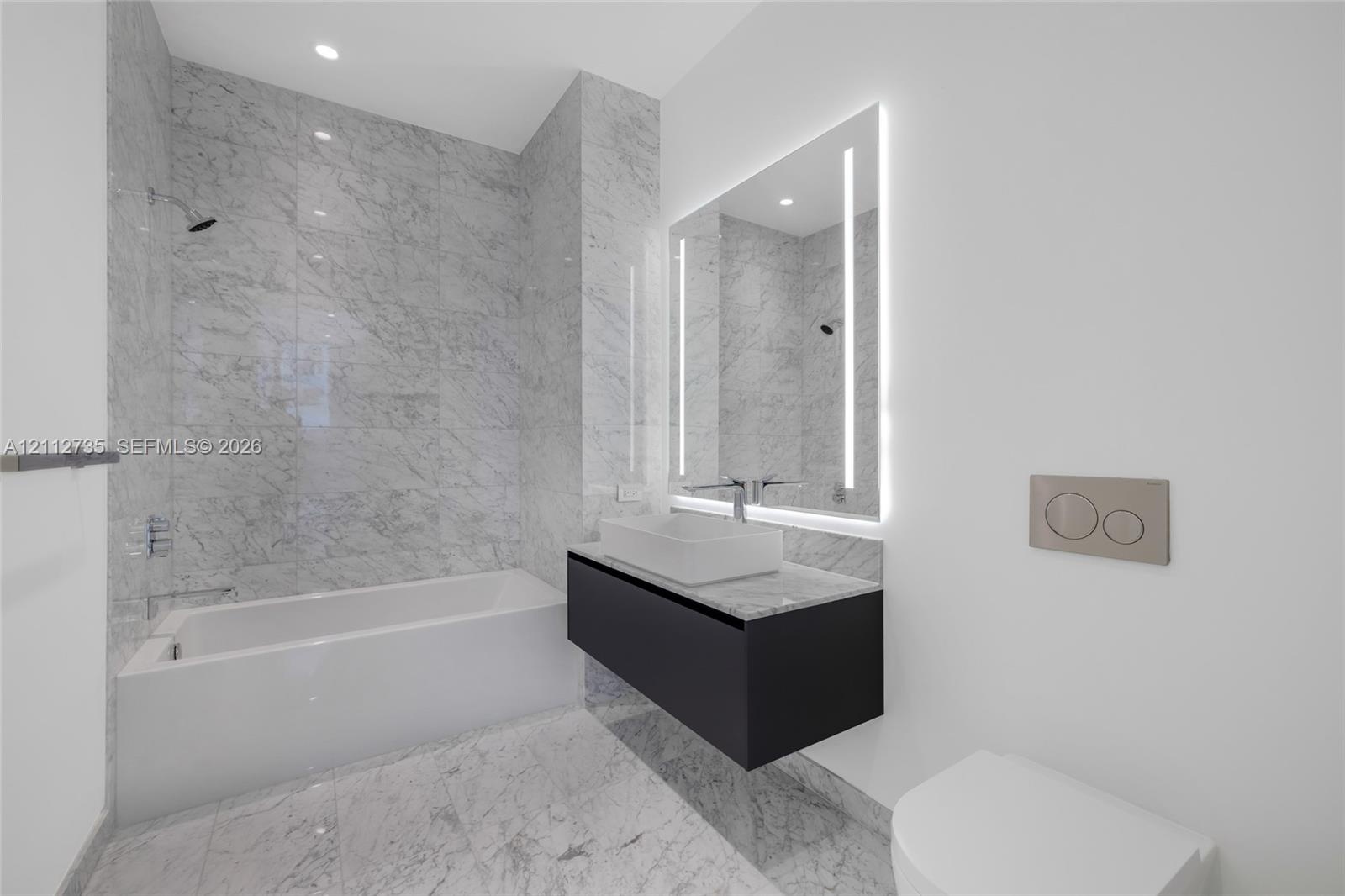 Full-height stone wall tiling, floating vanity with vessel sink, wall-mounted faucet, illuminated mirror, and wall-mounted dual-flush toilet