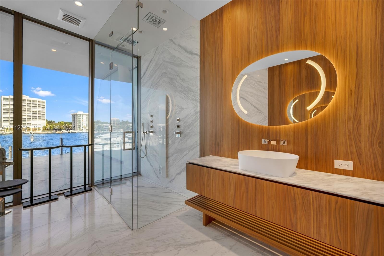 Floating wood-finish vanity with a stone countertop and vessel sink, illuminated oval mirror, walk-in glass-enclosed shower, stone-surround shower wal