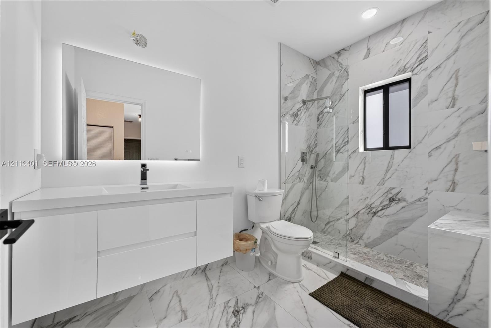 Floating white vanity with integrated sink, expansive LED-backlit mirror, floor-to-ceiling stone-look wall tiles, walk-in glass-enclosed shower, and m