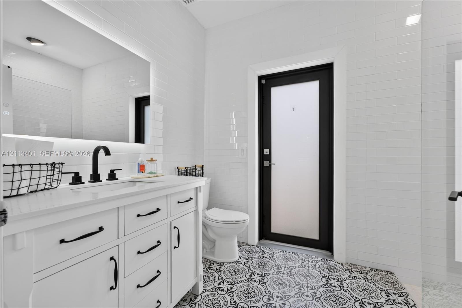 White vanity with a light-toned countertop, matte black hardware, integrated backsplash, backlit mirror, and patterned tile flooring