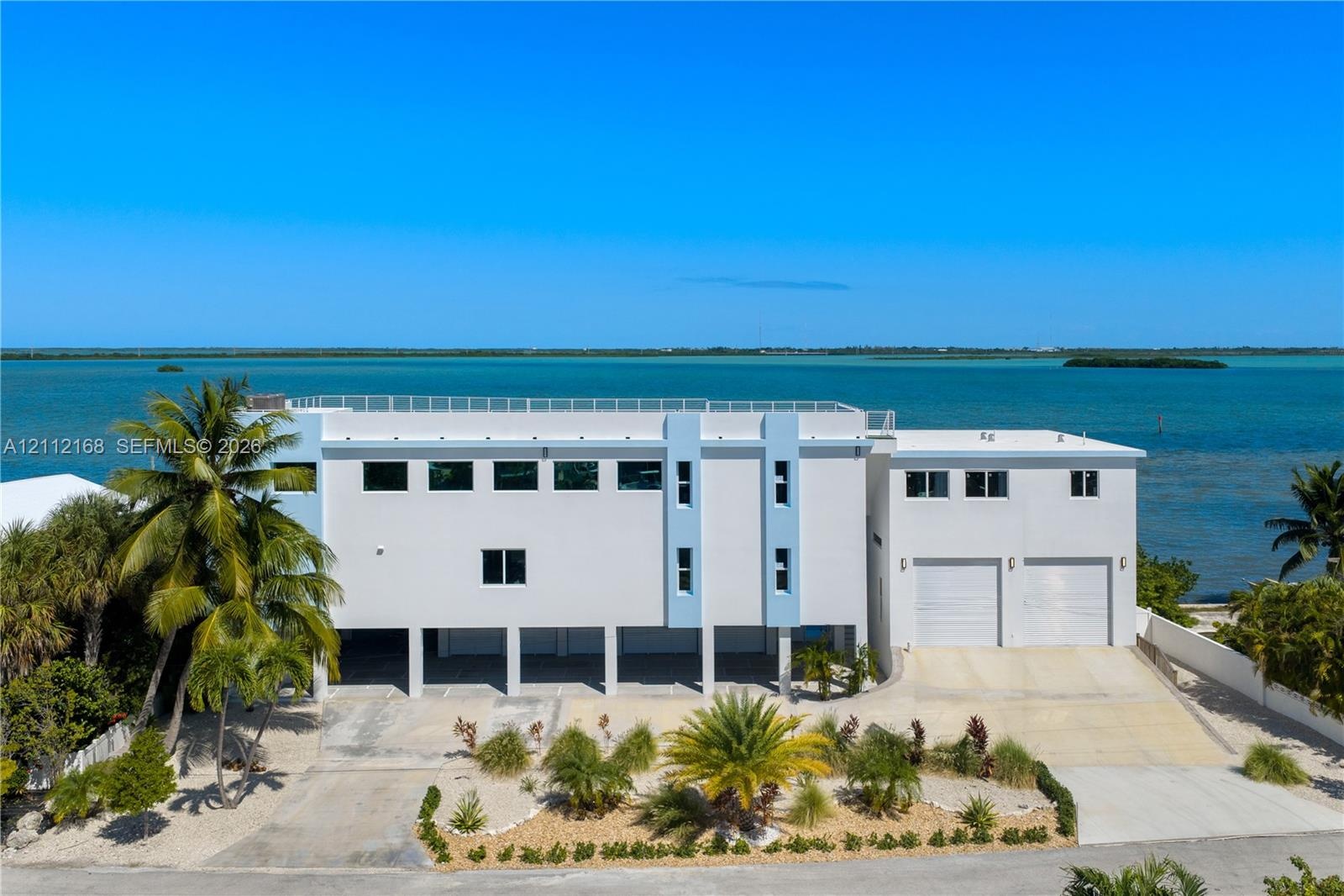 Multi-level coastal residence featuring an elevated structure, rooftop terrace with railing, dual garage bays, expansive concrete driveway, and direct