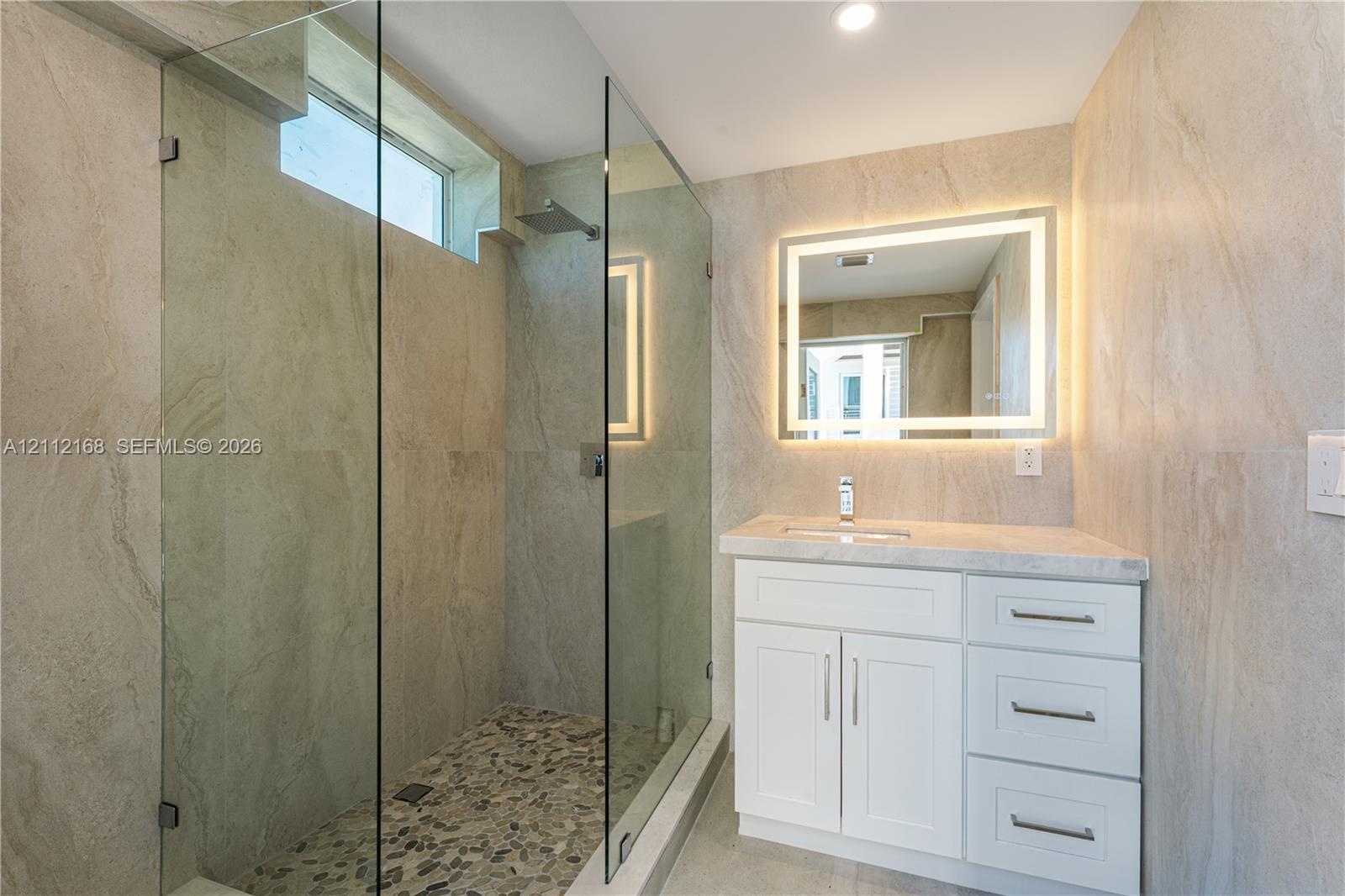 Walk-in shower with a glass enclosure, pebble tile floor, and rainfall showerhead, complemented by a white vanity with a stone countertop, integrated 