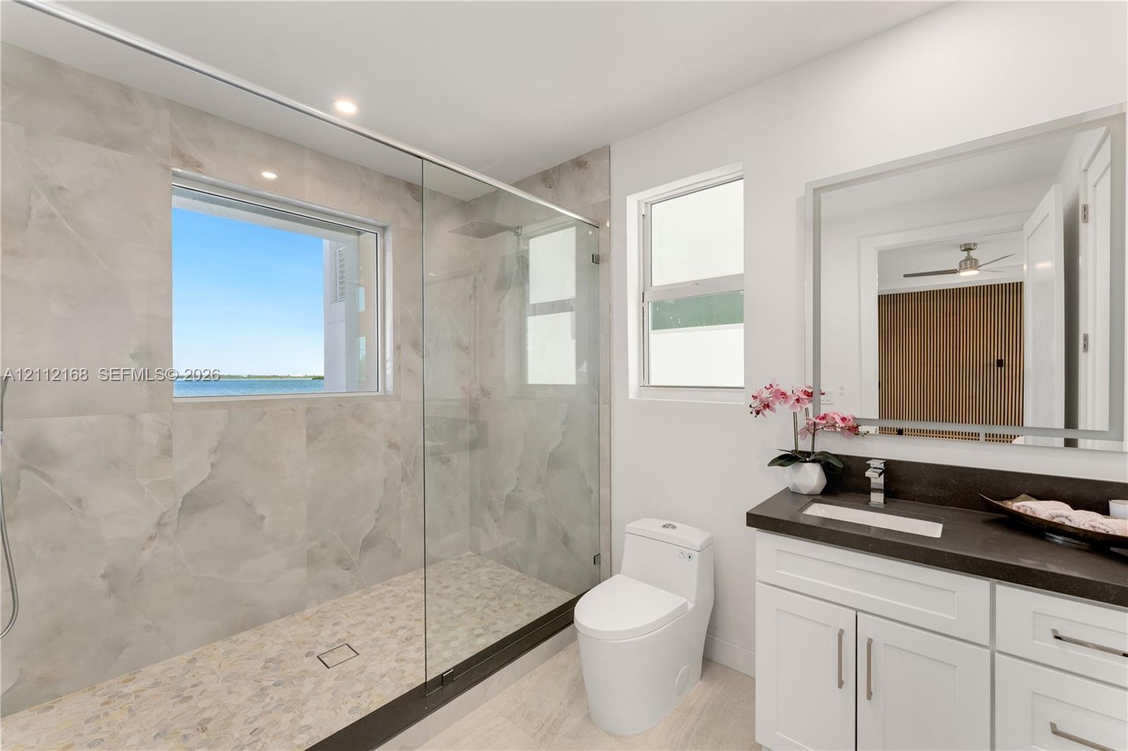 Bathroom featuring a walk-in shower with large-format wall tile and pebble-texture flooring, frameless glass enclosure, dual-pane window, and white ca
