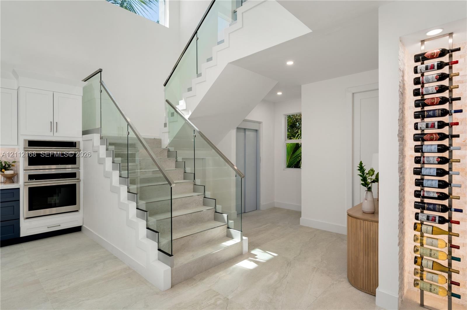 Open-riser staircase featuring a glass-panel railing, built-in stainless steel double wall ovens, recessed wine storage rack, and light-toned tile flo