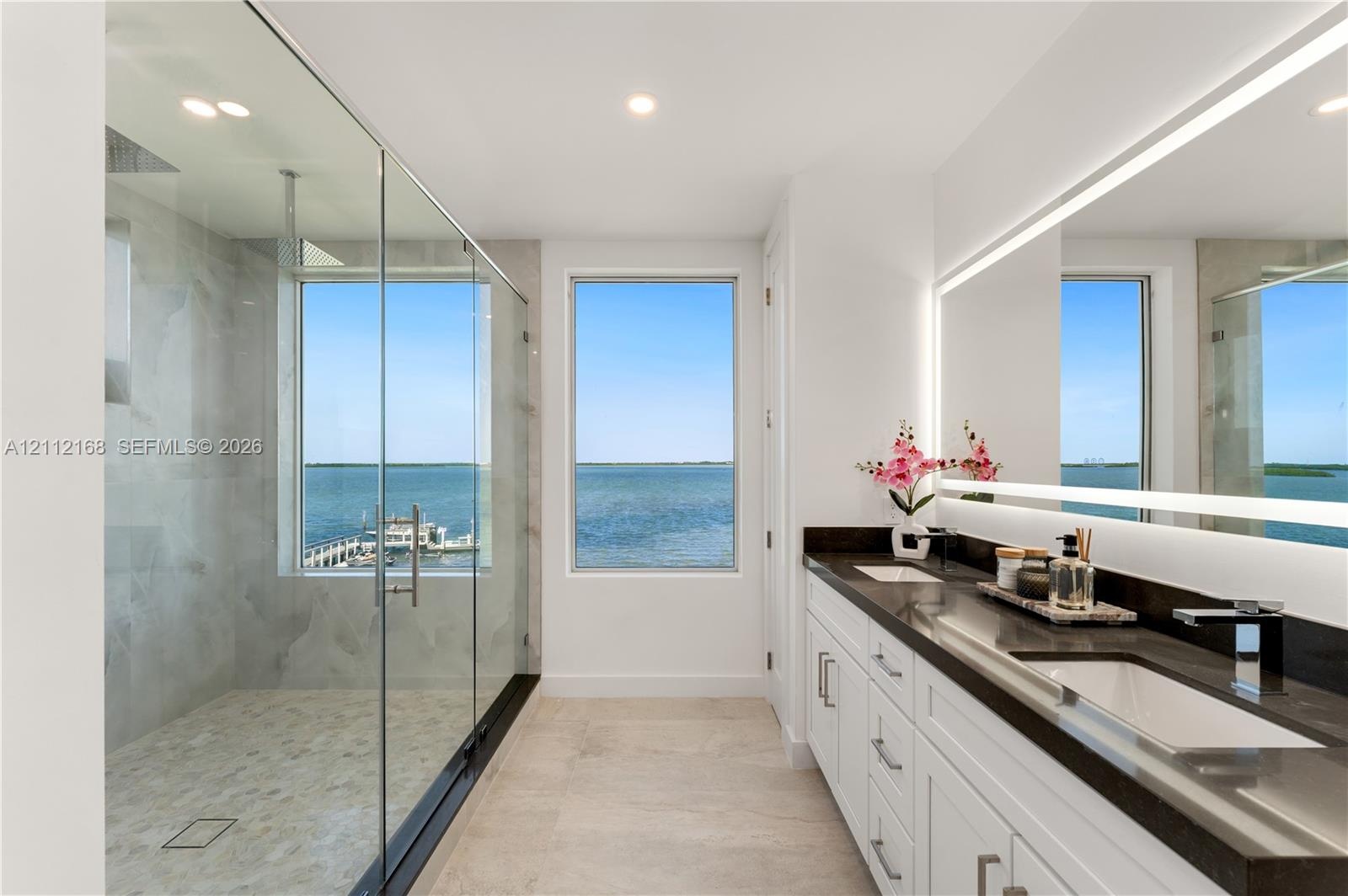 Dual-vanity bathroom featuring white cabinetry, dark stone countertops, a large frameless glass-enclosed shower with stone tile flooring, and a full-l