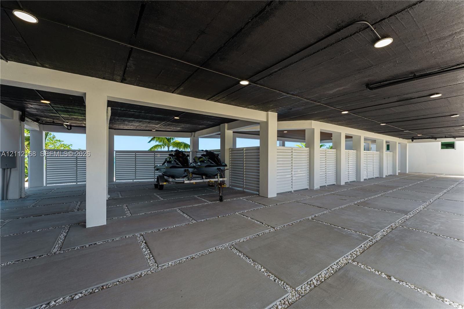 Covered parking area featuring large-format concrete pavers with stone aggregate borders, structural support pillars, white privacy screening, and rec
