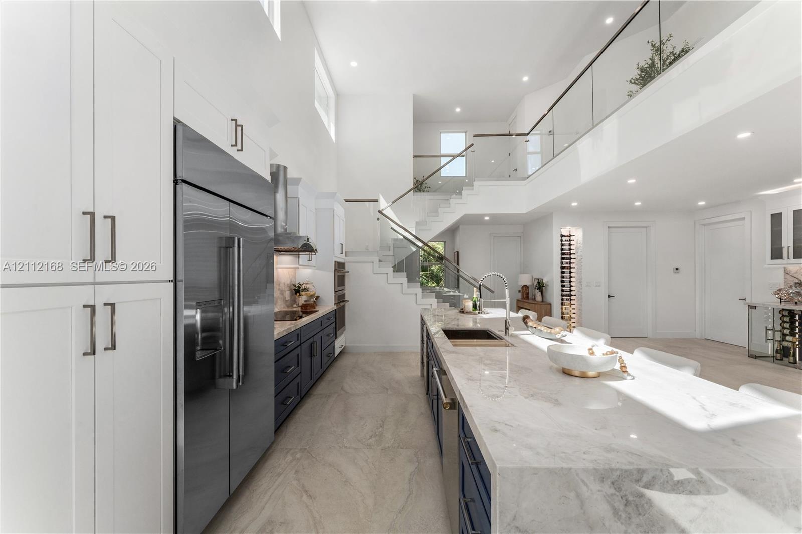 High-ceilinged kitchen featuring dual-tone cabinetry, a waterfall-edge stone island, recessed lighting, and a glass-panel staircase railing