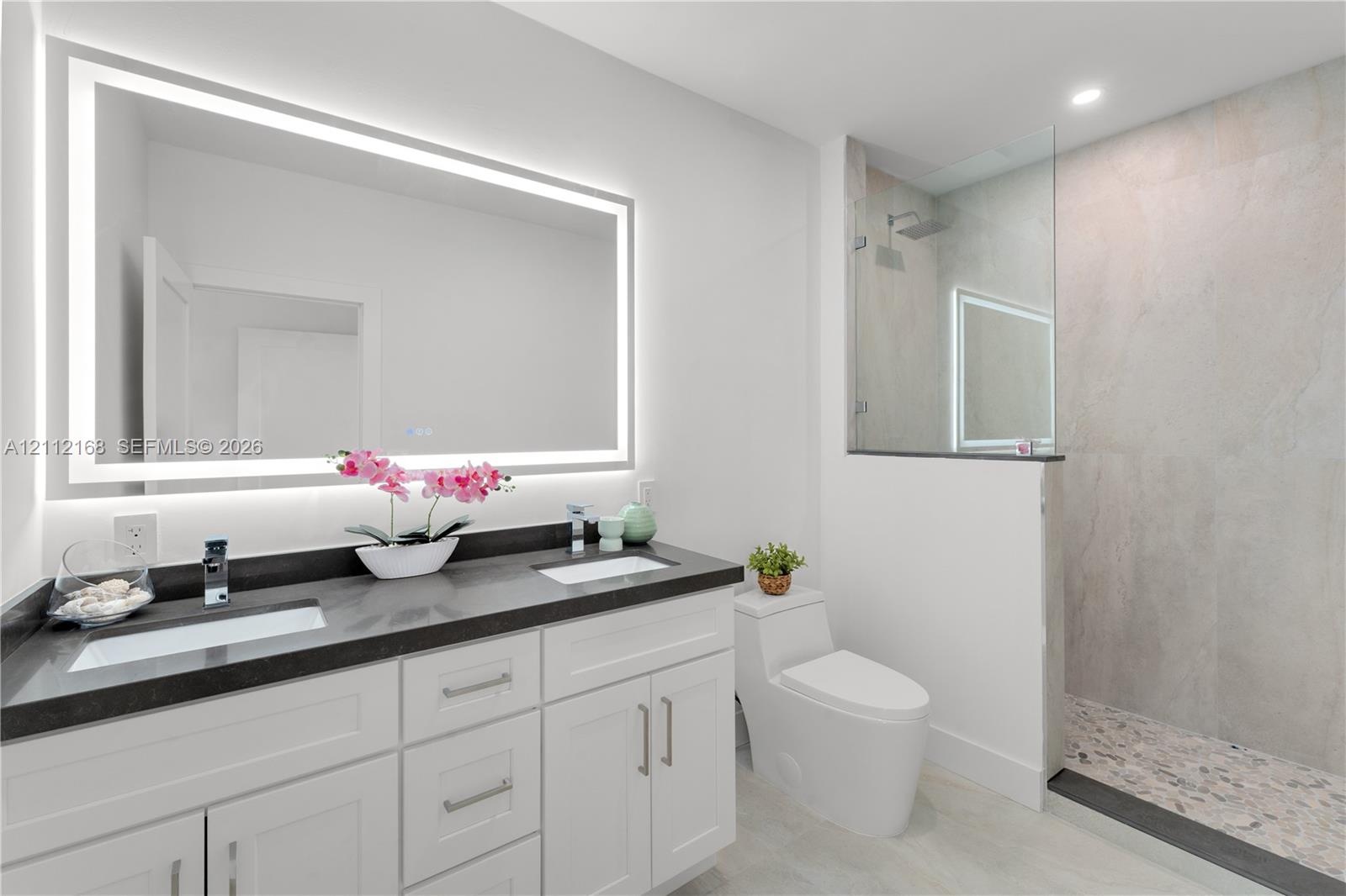Double vanity with dark countertops, integrated rectangular sinks, wall-to-wall LED backlit mirror, walk-in glass shower with stone-style tile surroun