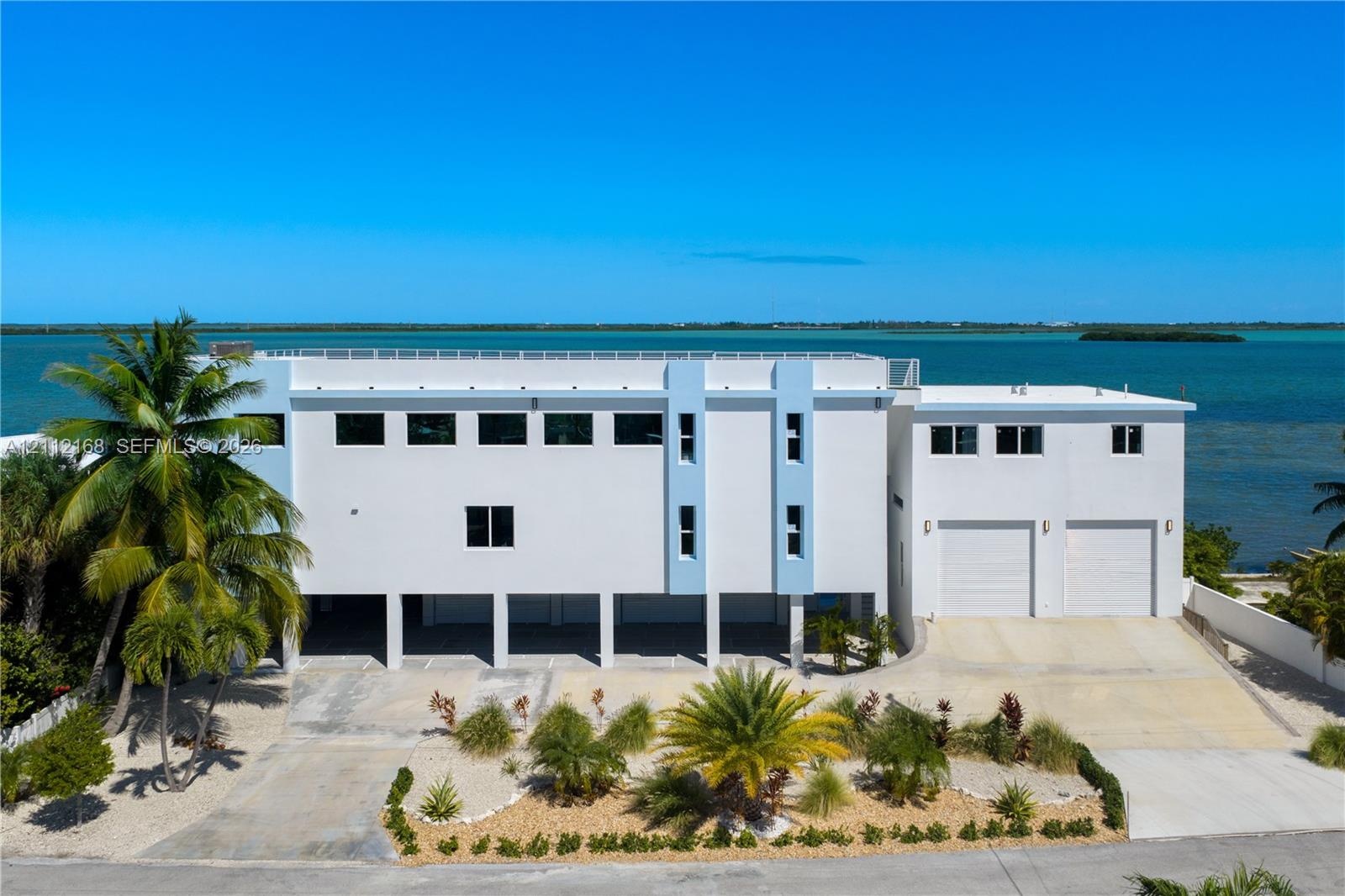 Multi-level contemporary exterior featuring a concrete facade, two-car garage, covered ground-level parking, rooftop railing, and waterfront orientati